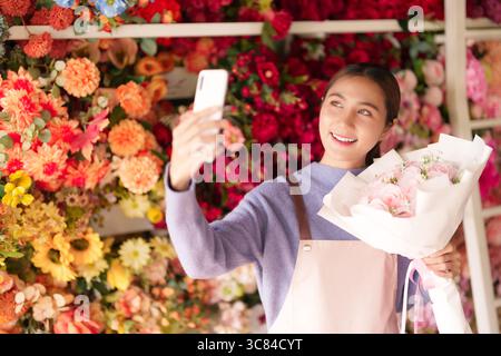 Il gioioso fiorista asiatico cattura un vibrante selfie che tiene fresche rose rosa all'interno della sua colorata boutique di fiori, perfetta per una promozione per piccole imprese Foto Stock