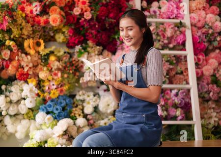 La fioraia asiatica adulta lavora in un vivace negozio di fiori che legge un taccuino che progetta composizioni floreali. Gestisce le attività quotidiane in modo allegro Foto Stock