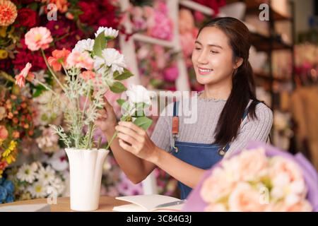 La fiorista asiatica sorridente donna adulta organizza sapientemente un vivace negozio di fiori. Le sue mani creano splendidi disegni floreali. Professionista creativo Foto Stock