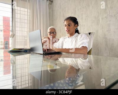 Nipote indiana che insegna alle donne anziane (nonna) a utilizzare il laptop a casa. Lavorare insieme per l'assegnazione scolastica utilizzando un laptop. Foto Stock