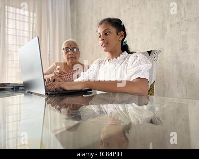 Nipote indiana che insegna alle donne anziane (nonna) a utilizzare il laptop a casa. Lavorare insieme per l'assegnazione scolastica utilizzando un laptop. Foto Stock