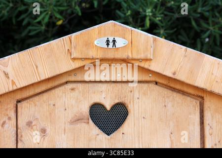 Un attico in legno con un cartello in metallo unisex per servizi igienici e un'apertura di ventilazione a forma di cuore sulla porta si erge contro il verde fogliame Foto Stock
