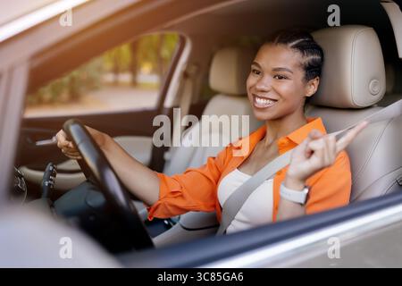 Sorridente giovane donna nera in auto che punta con gioia durante la guida Foto Stock