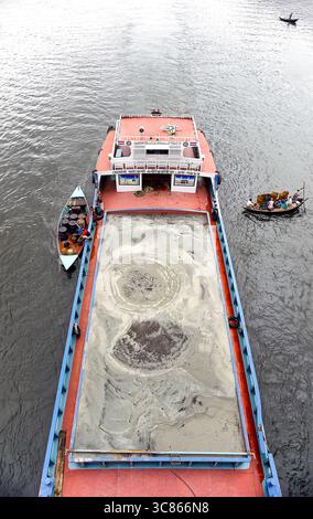 Una grande nave da carico che trasporta sabbia naviga in un fiume in Bangladesh, circondato da piccole barche di legno sulle calme acque marroni. Foto Stock