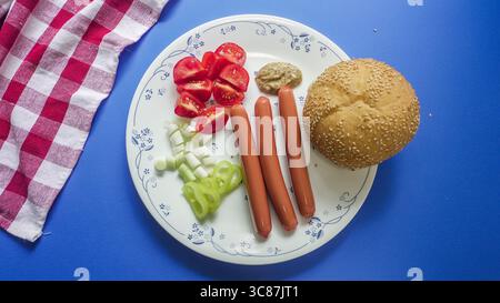Un piatto mostra tre hot dog insieme a pomodori a dadini, cetrioli e peperoni verdi. Un panino di semi di sesamo e senape completano il pasto, appoggiato contro a. Foto Stock