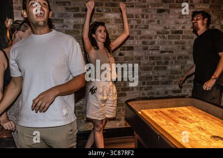 I giovani amici che celebrano una partita di shuffleboard vincono, mostrando eccitazione e cameratismo durante una divertente serata fuori in un bar. Foto Stock