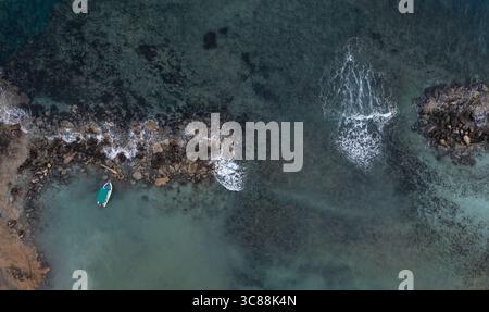 Tranquilla vista aerea con drone di una piccola barca da pesca ormeggiata vicino a una costa rocciosa, onde che si infrangono dolcemente, creando una tranquilla scena costiera Foto Stock