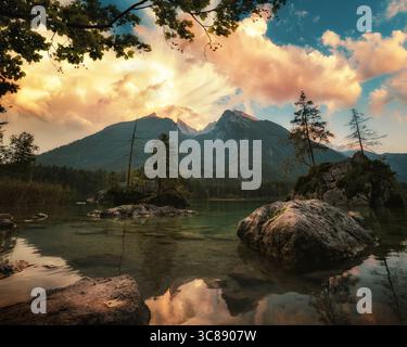 Nuvole dorate al tramonto sulle montagne Watzmann e sul lago Hintersee nelle Alpi tedesche. Paesaggio lungo la riva del lago con un cielo spettacolare. Foto Stock