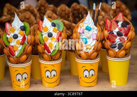 Una fila di colorate cialde di Hong Kong ripiene di panna montata, frutta fresca e dolci in un festival gastronomico cittadino. I dessert sono serviti in bicchieri di carta giallo brillante con illustrazioni sorridenti. Le varietà includono condimenti con fragole, kiwi, mirtilli, mango e marshmallow Foto Stock