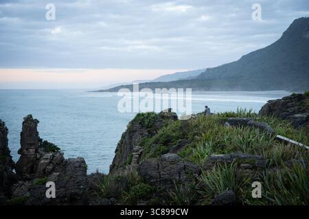Turista in un punto panoramico godendo di vedute sull'oceano e sul paesaggio costiero durante il tramonto, nuova Zelanda Foto Stock