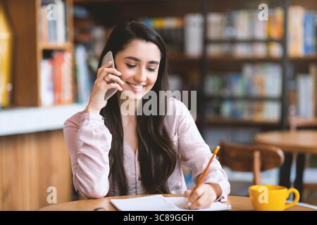 Caccia al lavoro. Donna araba felice che parla via smartphone con un rappresentante hr e prende appunti, seduta all'interno del bar Foto Stock