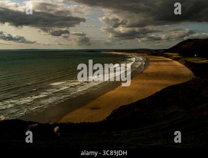Una vista panoramica di una spiaggia con onde che si infrangono sulla riva, sotto un cielo nuvoloso. La spiaggia è dorata, e ci sono delle persone sopra. In primo piano Foto Stock