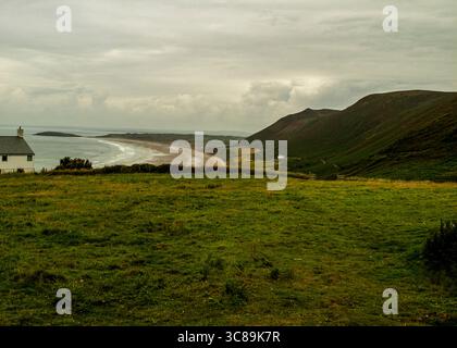 Un paesaggio panoramico caratterizzato da un campo erboso in primo piano, una spiaggia e l'oceano nel terreno centrale e una catena montuosa sullo sfondo. Un bianco Foto Stock