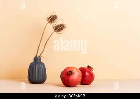 Cardo secco in vaso grigio e due melograni maturi su sfondo beige neutro. Natura morta autunnale minimalista, copia spazio. Foto Stock