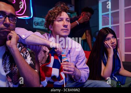 Gruppo di giovani amici multietnici adulti che guardano la partita sportiva in televisione, uomo caucasico che tiene la bandiera che sembra concentrata, donna asiatica che appare delusa, uomo nero sullo sfondo che copre il viso Foto Stock