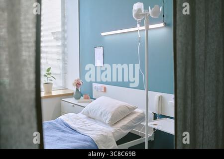 Luce solare che illumina il letto di ospedale vuoto con il supporto IV nella moderna stanza del paziente Foto Stock