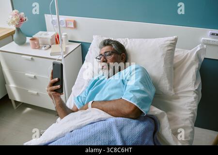 Uomo birazziale giacente nel letto di ospedale che utilizza lo smartphone per la comunicazione Foto Stock