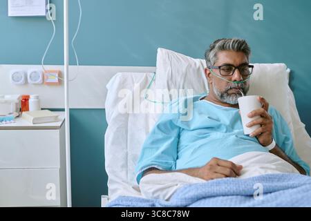 Uomo birazziale di mezza età che riposa nel letto di ospedale e beve da tazza Foto Stock