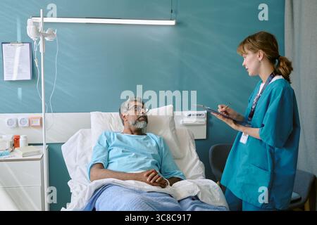 Uomo birazziale di mezza età che giace nel letto di ospedale che parla con il giovane infermiere adulto Foto Stock