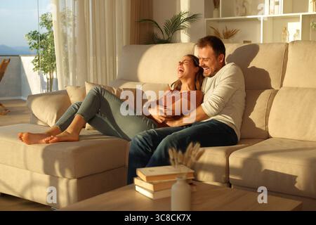 Coppia felice a casa con l'uomo che solletica la donna su un divano al tramonto Foto Stock