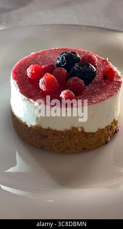 Una cheesecake cremosa si trova elegantemente su un piatto bianco, ricoperta da uno strato vibrante di frutti di bosco misti. La ricca consistenza del dessert e la frutta colorata garni Foto Stock