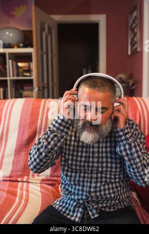 Uomo barbuto che ascolta la musica con le cuffie wireless in salotto Foto Stock