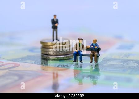 Miniaturgeschäftsmann auf Münzstapel hinter Rentnern auf Parkbank Symbolbild soziale Ungleichheit , Zwei Miniatur-Rentnerfiguren sitzen auf einer Bank vor einem Geschäftsmann auf einem Geldstapel. DAS Bild visualisiert symbolisch soziale Ungleichheit, Altersarmut und Vermögensverteilung im Ruhestand. *** Uomo d'affari in miniatura su un mucchio di monete dietro i pensionati su una panchina del parco immagine simbolica della disuguaglianza sociale , due figure in miniatura di pensionati siedono su una panchina di fronte a un uomo d'affari su una pila di soldi l'immagine simbolicamente visualizza disuguaglianza sociale, povertà in vecchiaia e ricchezza di Foto Stock