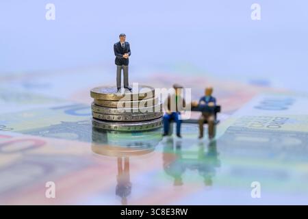 Miniaturgeschäftsmann auf Münzstapel hinter Rentnern auf Parkbank Symbolbild soziale Ungleichheit , Zwei Miniatur-Rentnerfiguren sitzen auf einer Bank vor einem Geschäftsmann auf einem Geldstapel. DAS Bild visualisiert symbolisch soziale Ungleichheit, Altersarmut und Vermögensverteilung im Ruhestand. *** Uomo d'affari in miniatura su un mucchio di monete dietro i pensionati su una panchina del parco immagine simbolica della disuguaglianza sociale , due figure in miniatura di pensionati siedono su una panchina di fronte a un uomo d'affari su una pila di soldi l'immagine simbolicamente visualizza disuguaglianza sociale, povertà in vecchiaia e ricchezza di Foto Stock