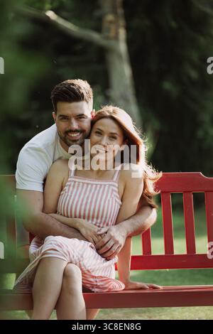 Felice giovane coppia in un parco. Uomo che abbraccia una bella donna da dietro mentre siede su una panchina rossa Foto Stock