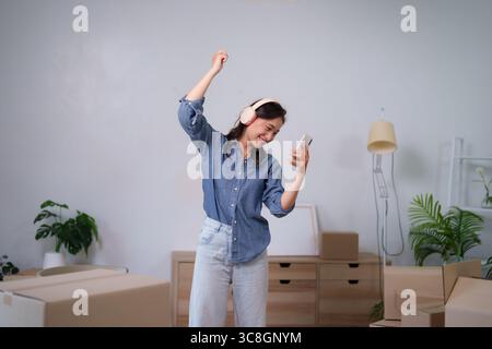 La donna asiatica felice ama la musica tramite cuffie e smartphone durante il giorno del movimento si rilassa nel suo nuovo appartamento circondato da scatole ed essenziale per la casa Foto Stock