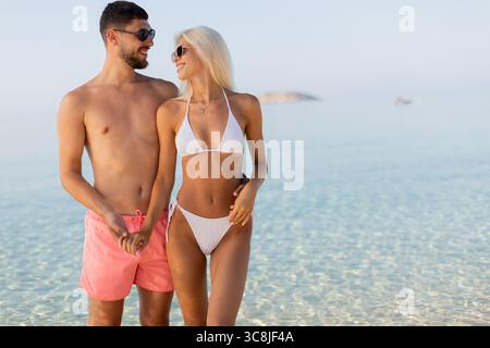 Una giovane coppia sta mano nella mano in una spiaggia tranquilla, sorridendo e godendosi la compagnia reciproca mentre il sole tramonta sulle dolci onde. Entrambi sono dres Foto Stock