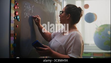 Insegnante donna che scrive sulla lavagna, parla con gli studenti, spiega il metodo di moltiplicazione dei numeri ai bambini delle scuole elementari durante la lezione di matematica. Corso di matematica in classe moderna e colorata Foto Stock