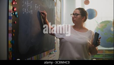 Insegnante donna che scrive sulla lavagna, parla con gli studenti, spiega il metodo di moltiplicazione dei numeri ai bambini delle scuole elementari durante la lezione di matematica. Corso di matematica in classe moderna e colorata Foto Stock