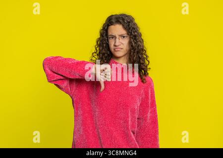 Non mi piace. Infelice ragazza caucasica che mostra un gesto di segno con il pollice giù, esprime malcontento disapprovazione, feedback negativo insoddisfatto. Bambino adolescente insoddisfatto in casa isolato su sfondo giallo Foto Stock