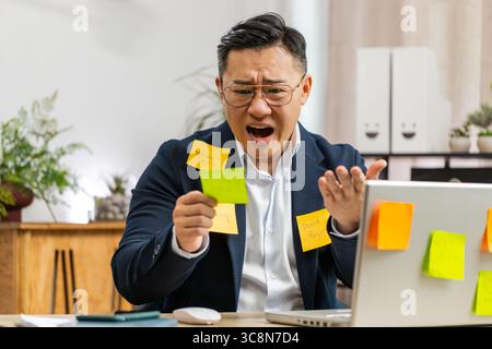 Uomo d'affari cinese stanco e stanco che lavora su un computer portatile in ufficio con molte iscrizioni sulle attività degli adesivi. L'uomo freelance che lavora da lavoro si sente tristemente noioso e ha lavorato troppo in multitasking. Attacco di panico, scadenza Foto Stock