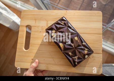 Human Hand tiene il cioccolato di Dubai su un tagliere di legno di fronte alla finestra. Splendido Sweet Cocoa Bar alla luce del giorno. Foto Stock