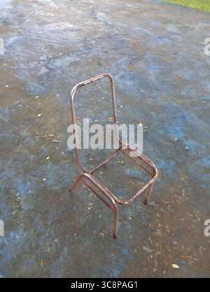 Il telaio della sedia in ferro, leggermente arrugginito, si erge su un pavimento di cemento blu con foglie asciutte sparse intorno ad esso. Foto Stock
