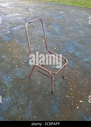 Il telaio della sedia in ferro, leggermente arrugginito, si erge su un pavimento di cemento blu con foglie asciutte sparse intorno ad esso. Foto Stock