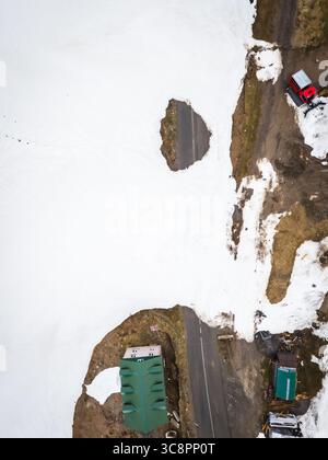 Vista aerea di una strada tortuosa che attraversa il passo innevato di Goderdzi, contrastando con macchie di terra marrone e piccoli edifici, Goderdzi, Adjara, Georgia. Foto Stock