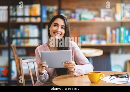 Donna araba felice seduta al bar, che usa un tablet digitale e beve caffè, sorride alla macchina fotografica, copia spazio Foto Stock