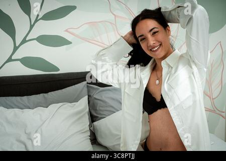 Donna sorridente che fissa i capelli mentre si siede sul letto in una camera da letto moderna con carta da parati floreale Foto Stock