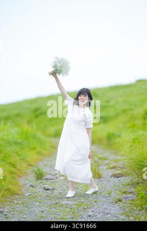 Una donna sui trent'anni con i capelli intermedi in un abito bianco si erge su un sentiero di montagna, sollevando un bouquet di respiro neonato nel cielo aperto, senza edifici. Foto Stock
