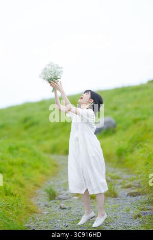Una donna sui trent'anni con i capelli intermedi in un abito bianco si erge su un sentiero di montagna, gettando delicatamente il respiro del bambino verso l'alto nel cielo aperto, senza edifici Foto Stock