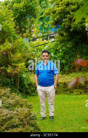 Uomo in piedi in un lussureggiante giardino verde con fiori colorati e denso fogliame, che indossa camicia blu e pantaloni crema. Foto Stock