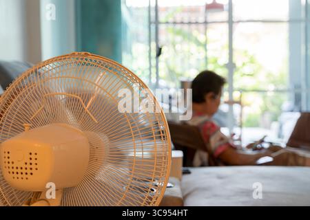 Primo piano di un ventilatore elettrico bianco in un accogliente soggiorno, che raffredda lo spazio mentre una donna anziana si rilassa sullo sfondo. Concetto di caldo estivo, ho Foto Stock