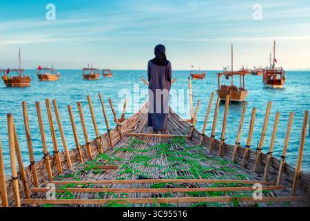 Le donne Abaya si trovano a prua di una tradizionale barca da pesca in legno, affacciata su un mare blu calmo punteggiato da barche simili. Foto Stock