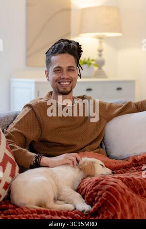 Uomo afroamericano sorridente e accarezzato cucciolo dormiente su coperta rossa nel soggiorno Foto Stock