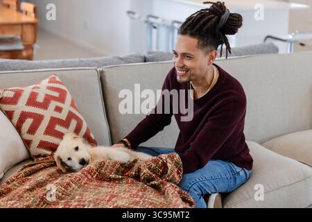 Uomo con dreadlock sorridente mentre coccola un piccolo cucciolo sotto una coperta intrecciata sul divano di casa Foto Stock