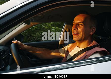 Uomo caucasico di mezza età che guida un'auto durante un viaggio in famiglia, sorridendo e guardando avanti con una mano sul volante, luce solare che illumina il viso, cintura di sicurezza allacciata Foto Stock