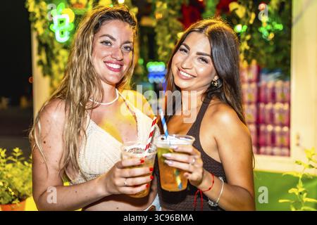 Due attraenti giovani donne sorridenti mentre tengono cocktail colorati ad una vivace festa in spiaggia, abbracciando completamente la gioia della vita notturna estiva Foto Stock